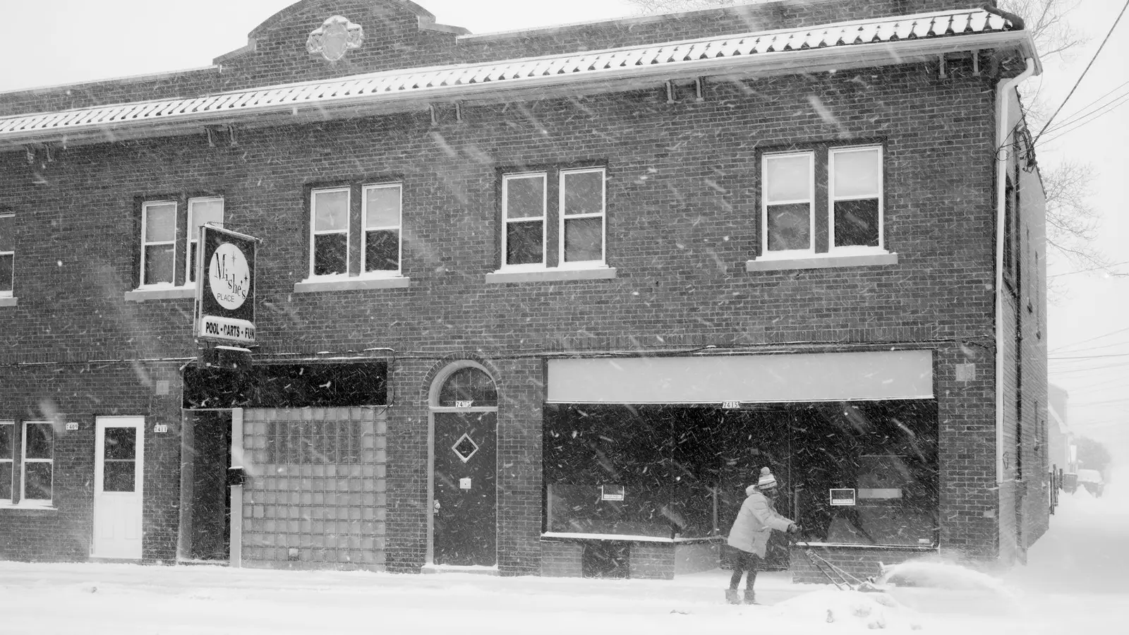 Person clearing a snow-covered sidewalk in front of a brick storefront building