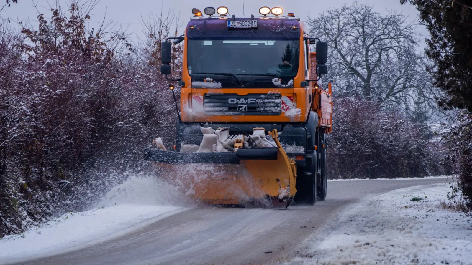 Orange plow truck clearing a tree-lined road during snowfall