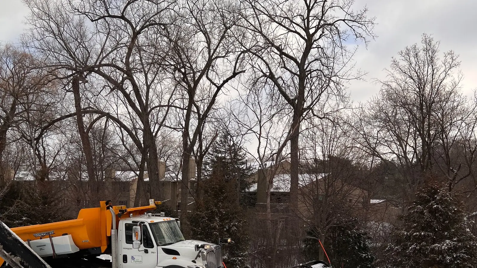 Plow truck with a yellow blade clearing a snowy road past bare trees