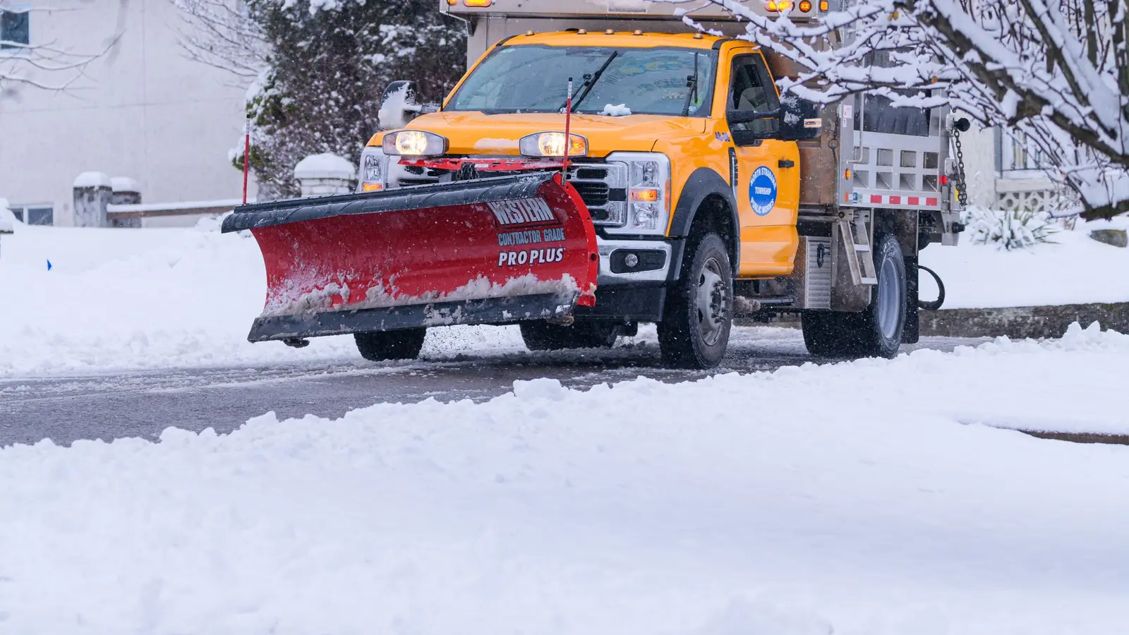 Orange salt truck with a red plow blade on a snowy road lined with frosted trees
