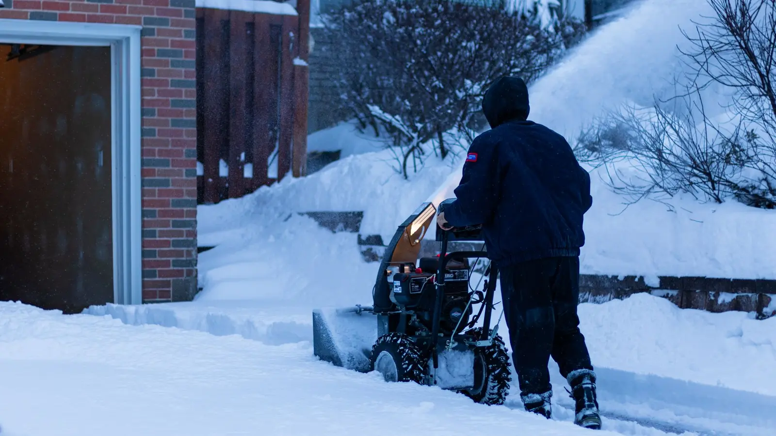 Person using a snowblower to clear a front walk beside a brick house